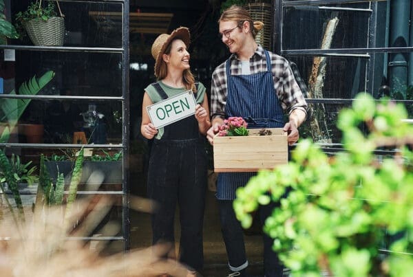 Contact - Smiling Young Business Owners Holding a Box of Produce and Open Sign Standing in Front of Their Shop