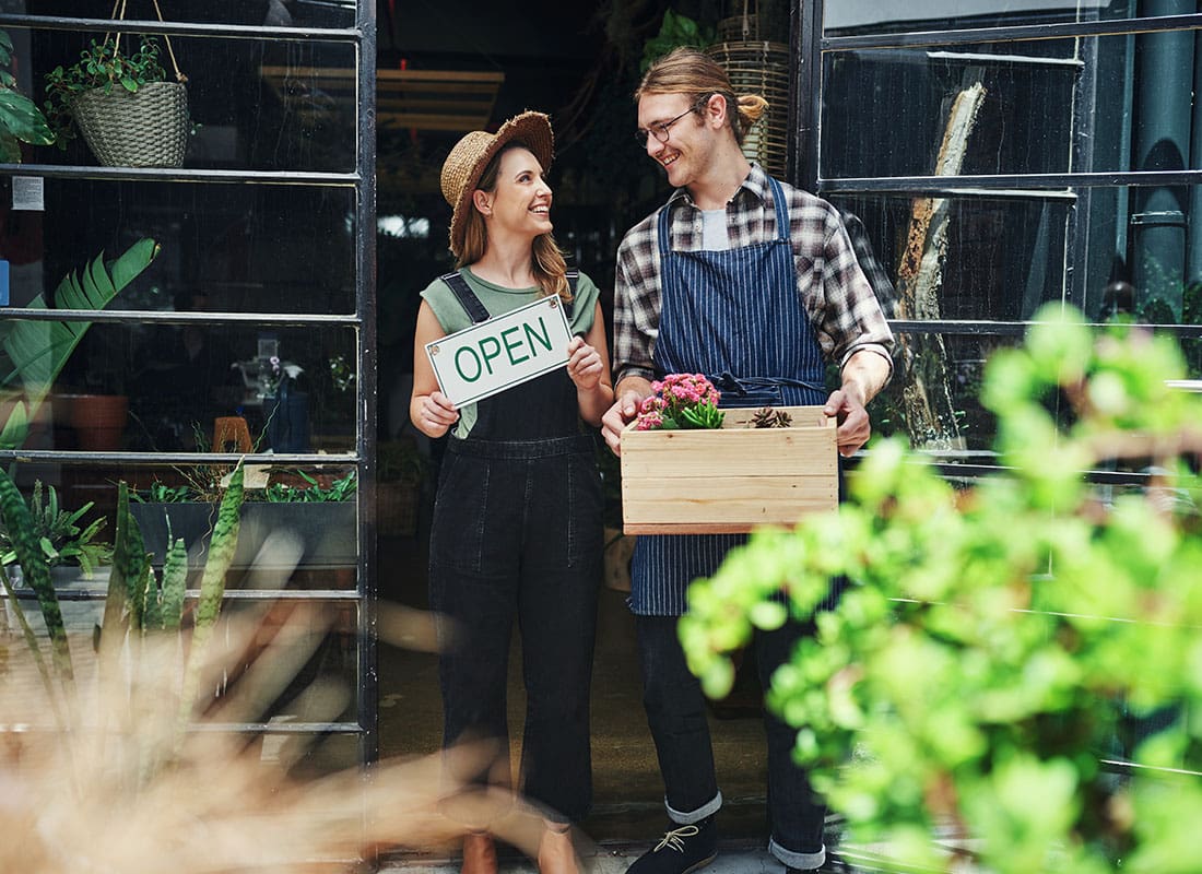 Contact - Smiling Young Business Owners Holding a Box of Produce and Open Sign Standing in Front of Their Shop